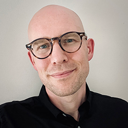 Nils smiles in a black button-up shirt and round tortoiseshell glasses in front of a light vignette background