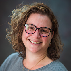 Johanna smiles in a gray shirt, black glasses, and a necklace in front of a charcoal-colored vignette background